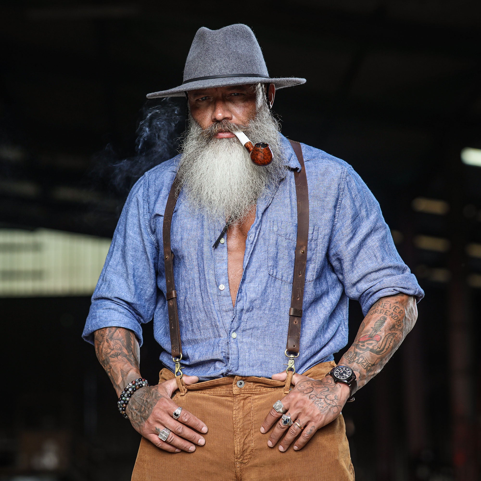 Man wearing Dark Brown Wiseguy Crazy Horse Suspenders over a denim shirt, styled with a pipe and hat in dark setting