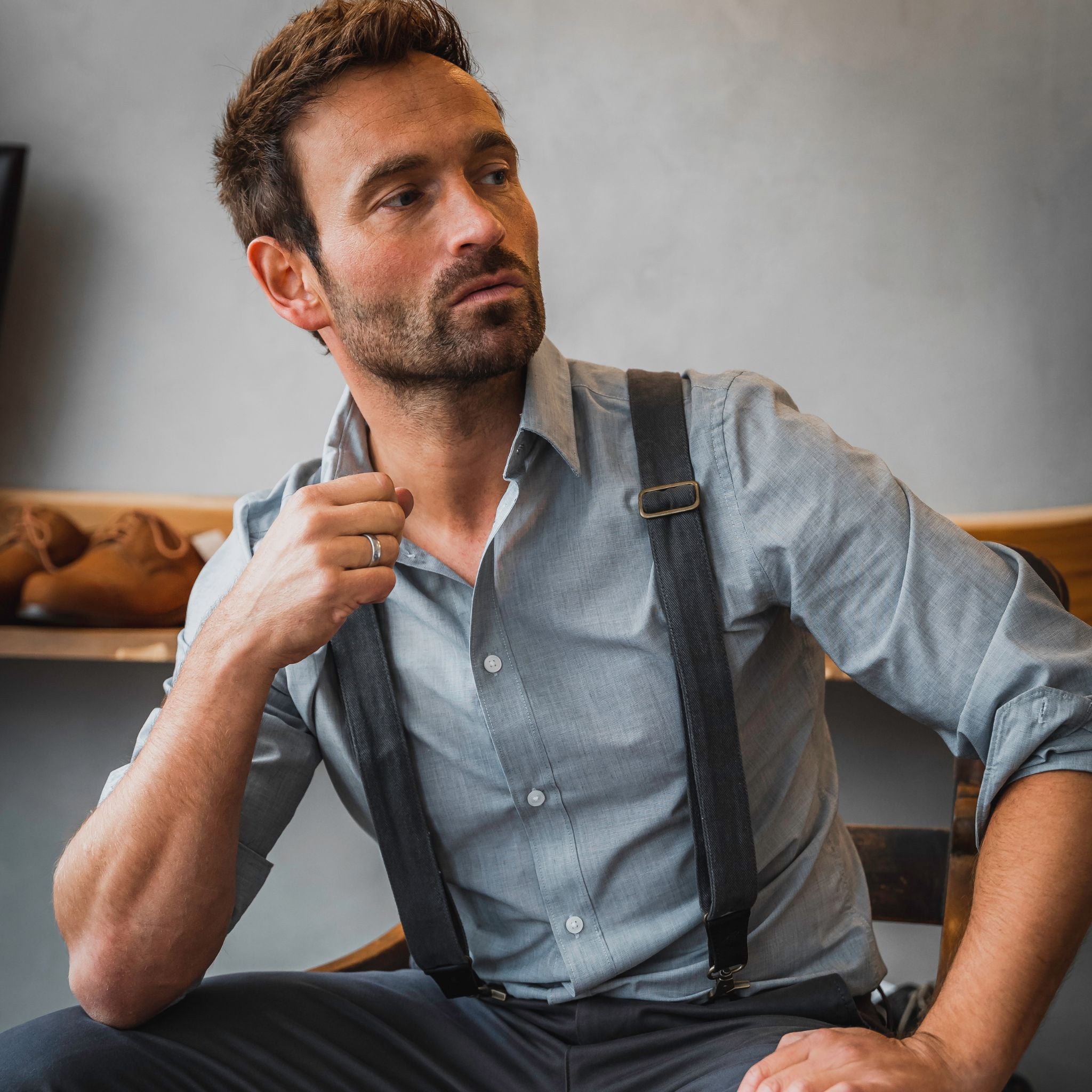 Man is sitting in chair, wearing a light grey shirt and dark grey pants and suspendersand 