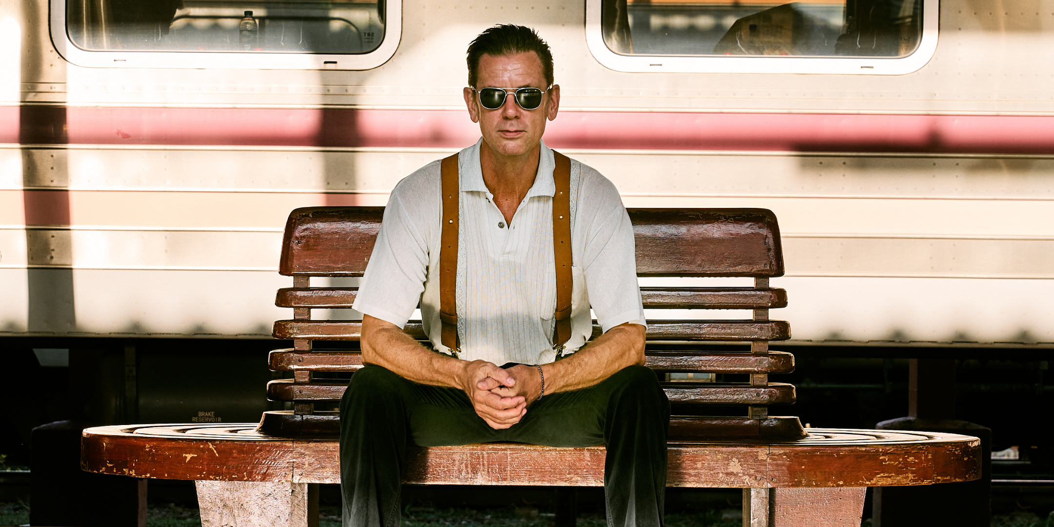 A man facing the camera, sitting on a bench at a train station. He is wearing aviator sunglasses and leather Wiseguy Original Crazy Horse Suspenders. There is a train behind him standing still. 