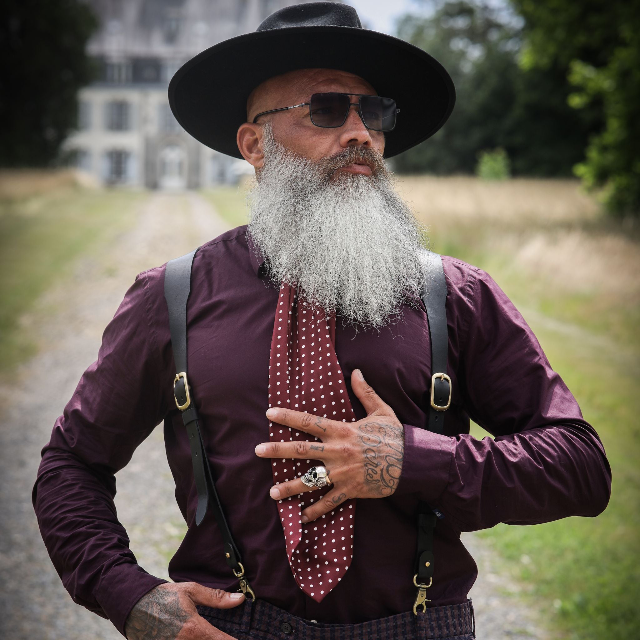 Man with white beard, hat, glasses and tie is wearing black leather suspenders and a burgundy shirt